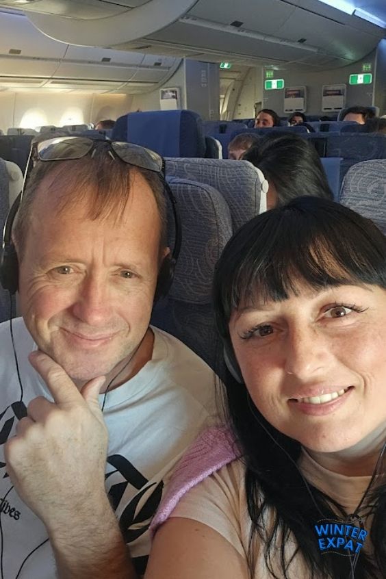 A close-up selfie of Ariana and Z smiling while seated inside the economy cabin of an Air China airplane. Z is wearing glasses on his head and a white graphic t-shirt, while Ariana is in the foreground wearing a light-colored top with a pink sweater over her shoulders. The WinterExpat logo is placed in the bottom right corner. Original photo by Ariana of WinterExpat.