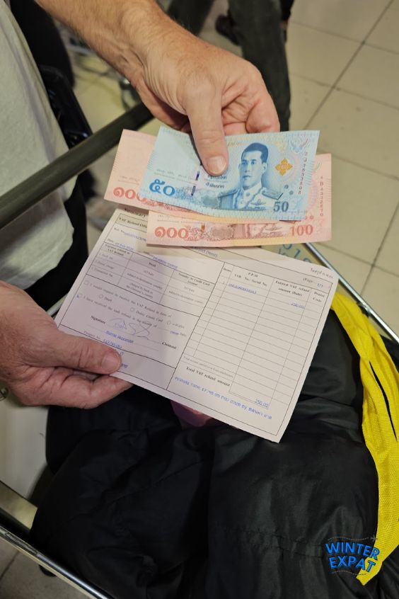 A close-up shot of a traveler's hands holding a filled-out Thai VAT refund application form on top of a black bag, with a 50 Baht note and a 100 Baht note resting on top of the document. Original photo by Ariana of WinterExpat.