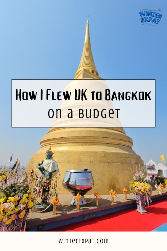 A tall vertical photograph featuring a massive, golden Buddhist chedi (stupa) against a clear blue sky. In the foreground, a teal and gold statue dressed in white stands next to a large silver offering bowl, surrounded by colorful flower arrangements on a red carpet. A translucent white text box overlay reads "How I Flew UK to Bangkok on a BUDGET". The WinterExpat logo is in the top right, and "WINTEREXPAT.COM" is printed at the bottom. Original photo by Ariana of WinterExpat.