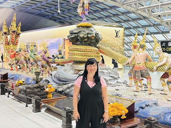 Ariana standing and smiling in front of the massive "Churning of the Milk Ocean" sculpture inside the Bangkok airport terminal. She is wearing a pink top and black jumpsuit, with the elaborate multi-figure Thai statue and a Louis Vuitton store visible in the background. Original photo by Ariana of WinterExpat.