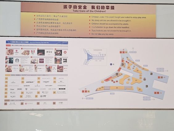 A close-up photograph of an informational terminal map at the Beijing airport. The top section of the sign features red and black text reading "孩子的安全 我们的牵挂 / Take Care of the Children!" alongside safety rules in both Chinese and English. Below it, an illustrated terminal layout map is displayed next to a directory of airport shops, cafes, and luxury brands. Original photo by Ariana of WinterExpat.