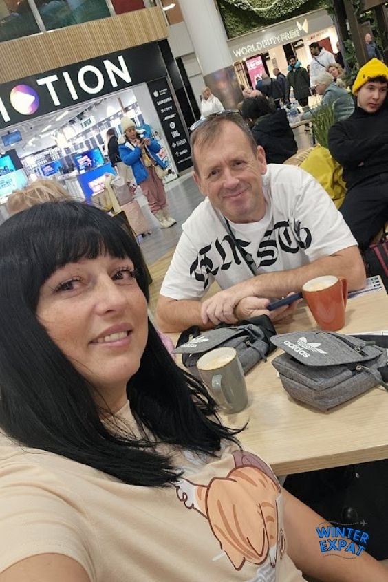 A selfie of Ariana and Z sitting at a light wooden table inside a busy airport terminal cafe. Ariana smiles in the foreground, while Z sits opposite her holding his phone next to an orange mug of coffee. In the background, airport shops like "InMotion" and "World Duty Free" are visible with other travelers walking by. The WinterExpat logo sits in the bottom right corner. Original photo by Ariana of WinterExpat.