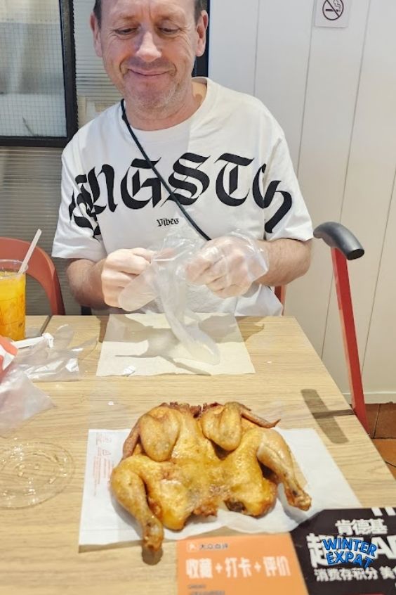 A photograph of Z sitting at a restaurant table, smiling as he puts on clear plastic gloves to eat. In the foreground on the wooden table, a whole roasted or fried chicken is served on parchment paper next to an orange card with Chinese text. The WinterExpat logo is visible in the bottom right corner. Original photo by Ariana of WinterExpat.
