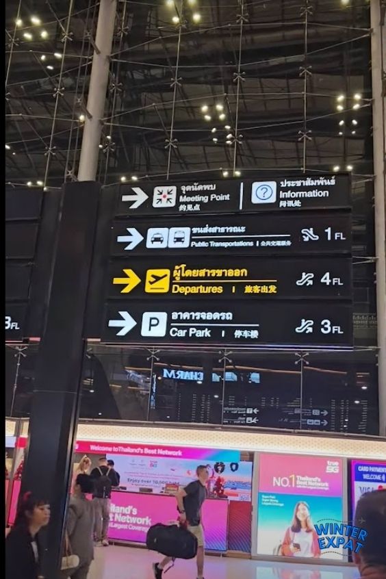 A photograph looking up at a large black airport directional sign inside a modern terminal with a high glass and steel ceiling structure. The sign displays directions with arrows and icons for the Meeting Point, Information, Public Transportations (1st floor), Departures (4th floor), and Car Park (3rd floor) in Thai, English, and Chinese. Colorful advertisements and travelers are visible at the bottom, with the WinterExpat logo in the lower right. Original photo by Ariana of WinterExpat.