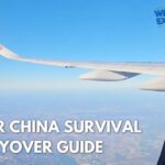 A view looking out of an airplane window showing the wing of an Air China aircraft flying high over land under a clear blue sky. The WinterExpat logo is visible in the top right corner, and the bottom left features the bold text overlay: "AIR CHINA SURVIVAL LAYOVER GUIDE". Original photo by Ariana of WinterExpat.