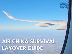 A view looking out of an airplane window showing the wing of an Air China aircraft flying high over land under a clear blue sky. The WinterExpat logo is visible in the top right corner, and the bottom left features the bold text overlay: "AIR CHINA SURVIVAL LAYOVER GUIDE". Original photo by Ariana of WinterExpat.