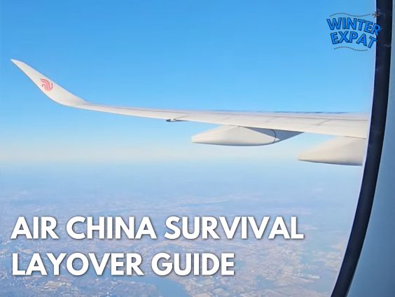 A view looking out of an airplane window showing the wing of an Air China aircraft flying high over land under a clear blue sky. The WinterExpat logo is visible in the top right corner, and the bottom left features the bold text overlay: "AIR CHINA SURVIVAL LAYOVER GUIDE". Original photo by Ariana of WinterExpat.
