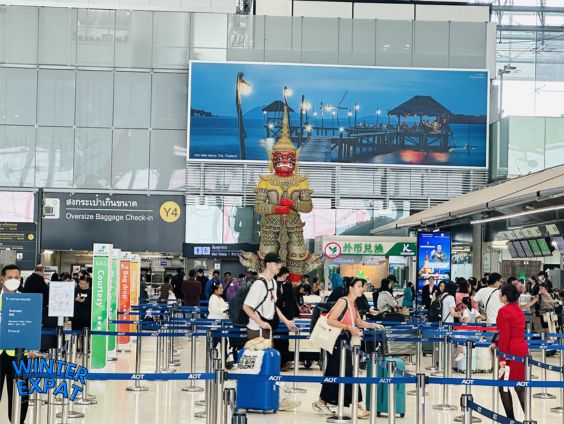 A bustling view of the oversized baggage check-in area at Suvarnabhumi Airport terminal. Travelers with luggage stand behind blue AOT queue ropes, with a tall, menacing red Yaksha guardian giant statue standing watch in the background beneath a large billboard of a Thai beach. Original photo by Ariana of WinterExpat.