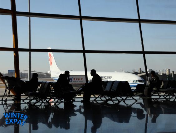 A silhouette photograph looking out through a large, floor-to-ceiling airport terminal window at an Air China airplane parked on the tarmac. Inside, the dark silhouettes of travelers are seen sitting in a row of terminal seats, with their shapes softly reflected on the polished airport floor. The WinterExpat logo is positioned in the bottom left corner. Original photo by Ariana of WinterExpat.