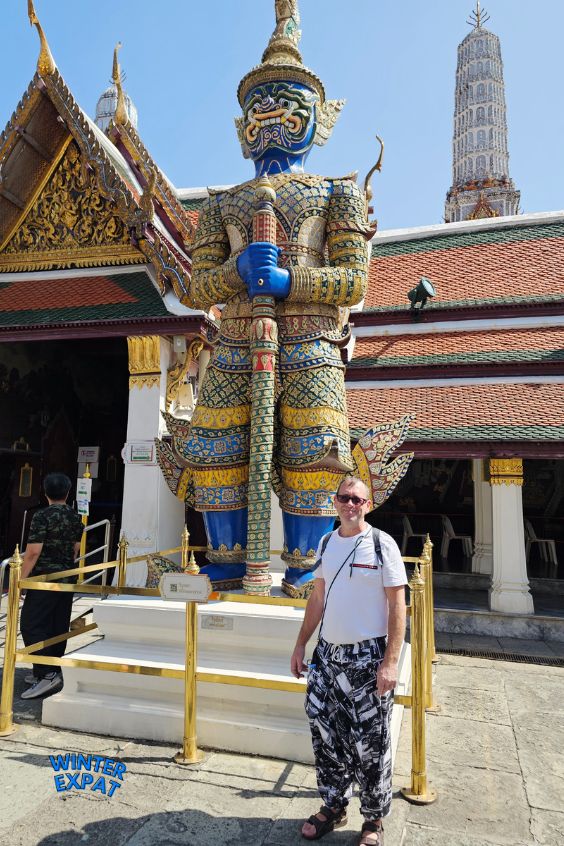 Z is standing and smiling in front of a giant, colorful demon guardian statue with a blue face at the Grand Palace in Bangkok. Original photo by Ariana of WinterExpat.