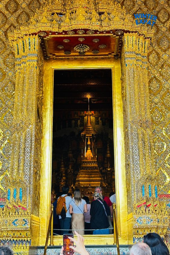 A close-up view of a large, highly detailed golden doorway at the Grand Palace in Bangkok. Through the open entrance, tourists are looking inside at a glowing golden structure or altar in the dark room. Original photo by Ariana of WinterExpat. 