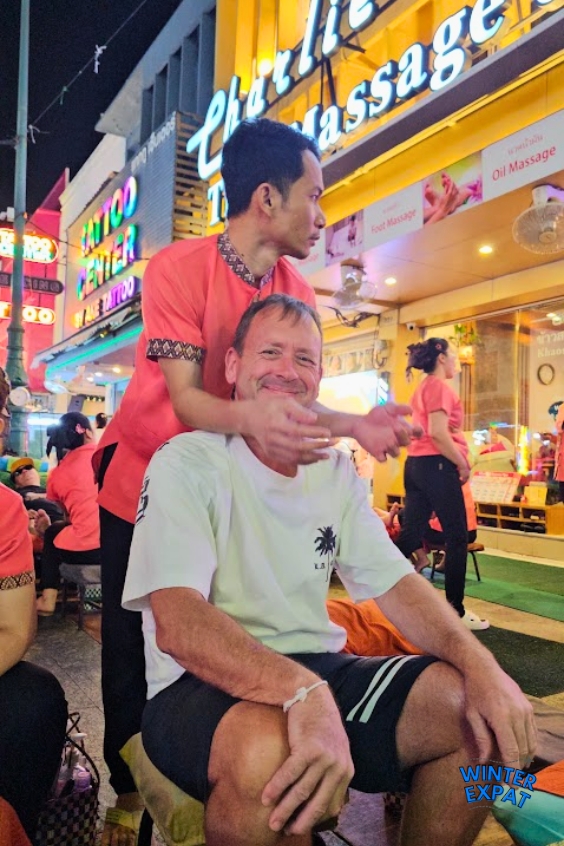 Z enjoys a shoulder and neck massage from a therapist at a busy street massage shop on Khaosan Road, surrounded by colorful lights and evening crowds. Original photo by Ariana of WinterExpat.
