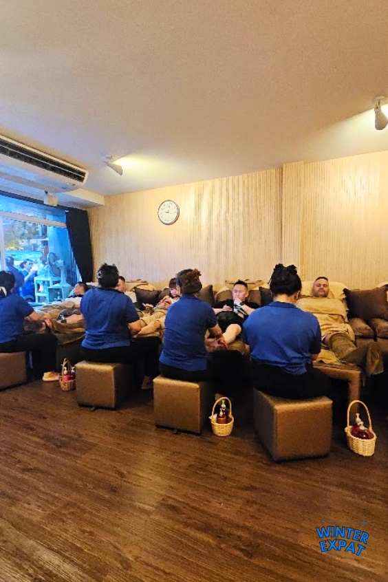 Guests relax on reclining chairs while therapists provide traditional Thai foot massages in a spacious massage room at Lavender Massage in Bangkok. Original photo by Ariana of WinterExpat.