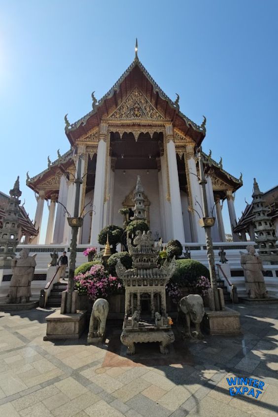 Front view of the main ordination hall at Wat Suthat with decorative stone pagodas, elephant statues, flowering gardens, and intricate Thai architecture. Original photo by Ariana of WinterExpat.