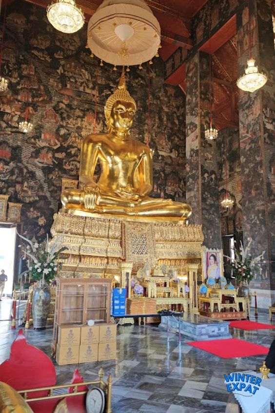 Large golden Buddha statue inside the main prayer hall of Wat Suthat, surrounded by ornate murals, chandeliers, and traditional Thai decorations. Original photo by Ariana of WinterExpat.