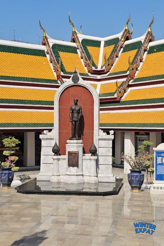 Bronze statue in the courtyard of Wat Suthat with the temple's yellow and green tiled roof rising behind it in Bangkok. Original photo by Ariana of WinterExpat.