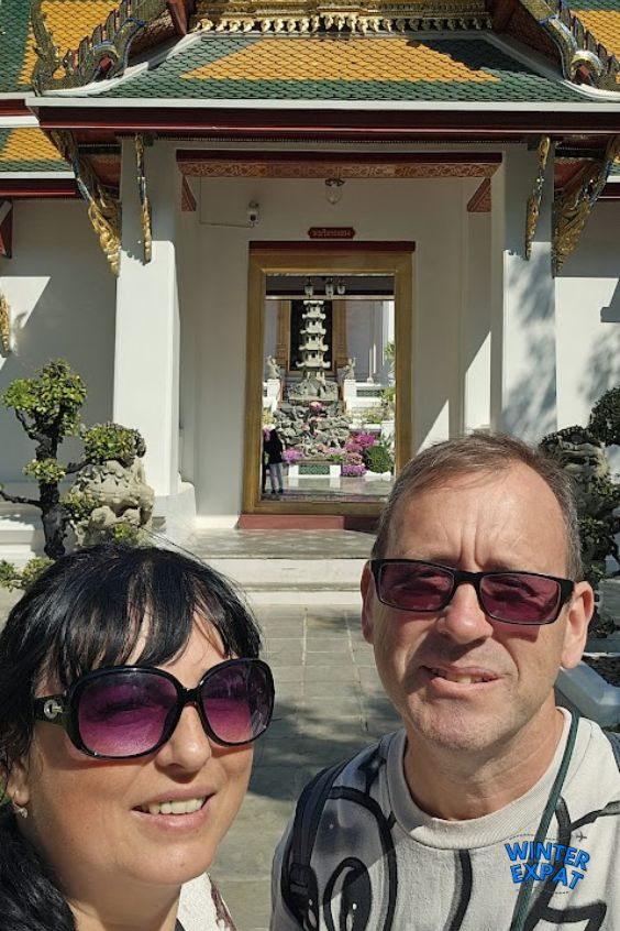 Ariana and Z standing in front of a temple entrance at Wat Suthat in Bangkok, with traditional Thai architecture and landscaped gardens behind them. Original photo by Ariana of WinterExpat.