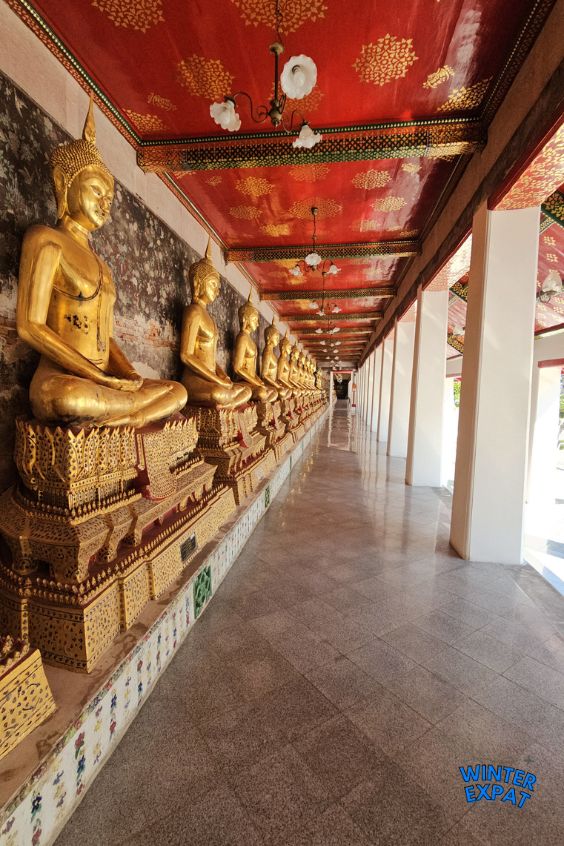 Long corridor at Wat Suthat lined with seated golden Buddha statues beneath a red and gold decorated ceiling in Bangkok. Original photo by Ariana of WinterExpat.