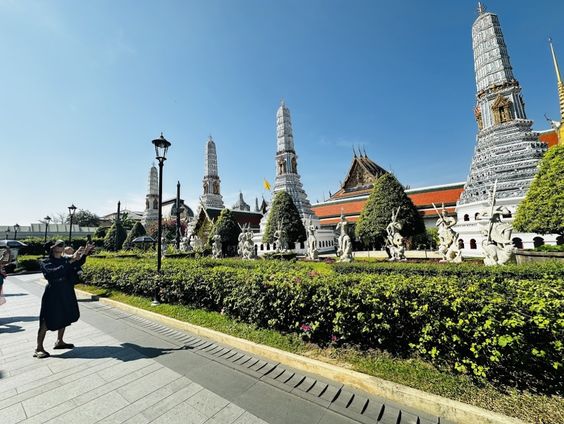 People are standing on a paved walkway, holding up their phones to take a photo of the Grand Palace grounds in Bangkok. In the background, there is a green hedge, several tall white towers, and statues under a bright blue sky. Original photo by Ariana of WinterExpat.