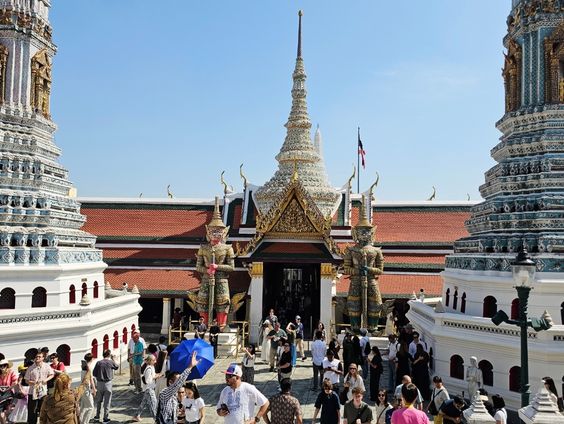 A busy courtyard at the Grand Palace in Bangkok, filled with tourists walking between large white towers. In the background, two tall demon guardian statues stand at the entrance of a building with an orange roof. Original photo by Ariana of WinterExpat.