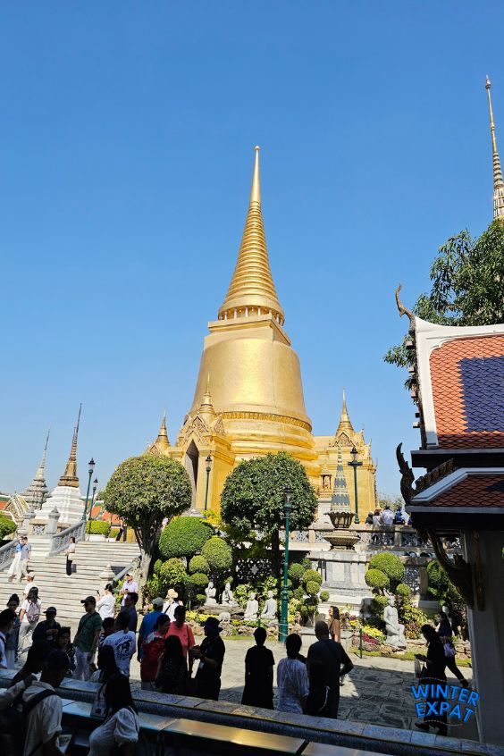 A tall, golden bell-shaped tower stands high under a bright blue sky at the Grand Palace in Bangkok. Below it, many tourists are walking around the courtyard near green trees and temple steps. Original photo by Ariana of WinterExpat. 