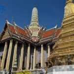 A beautiful gold and colorful temple at the Grand Palace in Bangkok under a bright blue sky. You can see tourists walking on the path below the buildings. Original photo by Ariana of WinterExpat.