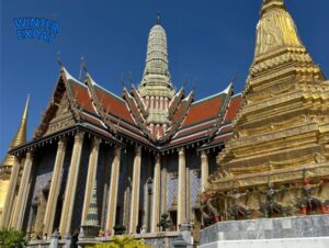 A beautiful gold and colorful temple at the Grand Palace in Bangkok under a bright blue sky. You can see tourists walking on the path below the buildings. Original photo by Ariana of WinterExpat.