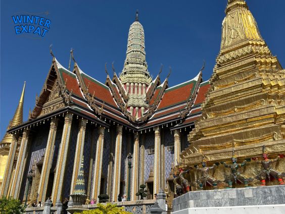 A beautiful gold and colorful temple at the Grand Palace in Bangkok under a bright blue sky. You can see tourists walking on the path below the buildings. Original photo by Ariana of WinterExpat.