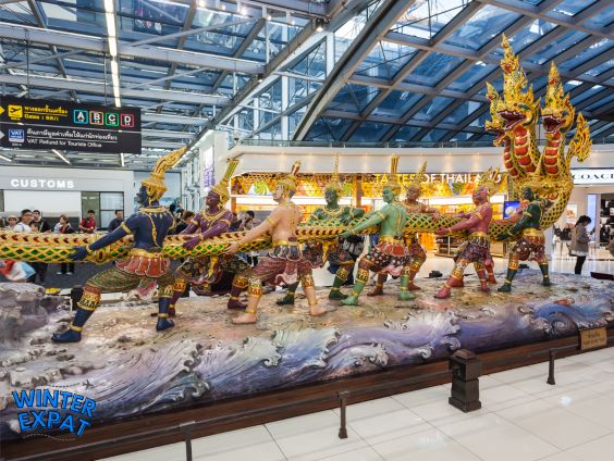 A wide graphic showing the large, colorful "Churning of the Milk Ocean" statue inside Suvarnabhumi Airport in Bangkok. Multiple traditional Thai figures pull a giant naga serpent. Large text overlay reads "SUVARNABHUMI ARRIVAL GUIDE" across the top, with a WinterExpat logo in the bottom left corner. Original photo by Ariana of WinterExpat.