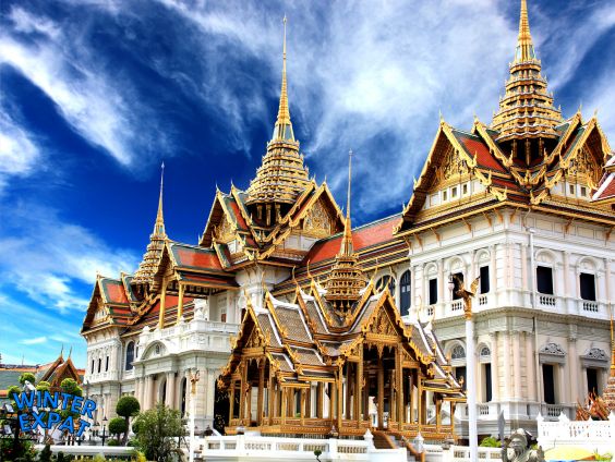 A beautiful view of the Chakri Maha Prasat Throne Hall at the Grand Palace in Bangkok under a bright blue sky with fluffy white clouds. The grand building mixes European style with golden traditional Thai roofs and sharp spires. Original photo by Ariana of WinterExpat.