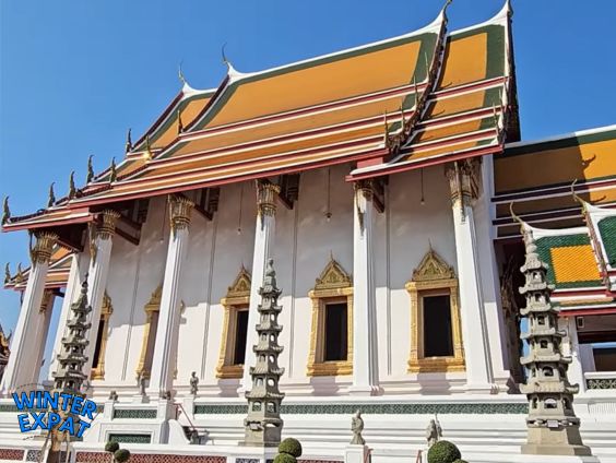 Side view of Wat Suthat's white temple building with ornate golden windows, traditional Thai roof, and decorative stone pagodas in Bangkok. Original photo by Ariana of WinterExpat.