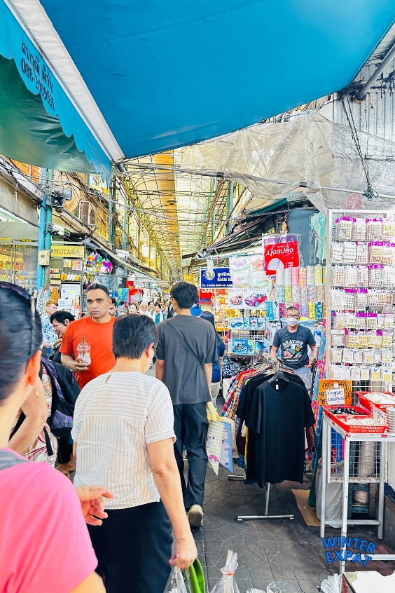 Crowds of shoppers walking through a busy covered alleyway lined with colorful clothing racks, accessories, and souvenir displays at Sampheng Market. Original photo by Ariana of WinterExpat.