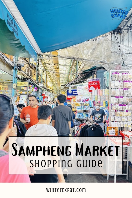 Crowded covered alleyway inside Sampheng Market lined with hanging goods, wholesale souvenir displays, and shoppers, with text overlay reading Sampheng Market Shopping Guide. Original photo by Ariana of WinterExpat.