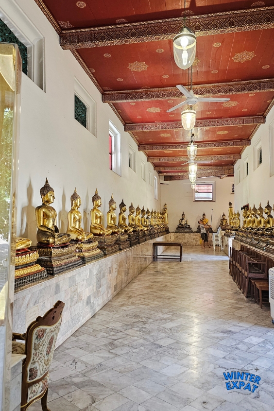 Long hallway inside Wat Pho lined with dozens of golden seated Buddha statues beneath a richly decorated red ceiling. Original photo by Ariana of WinterExpat.
