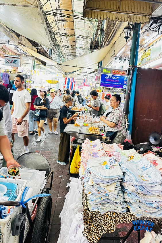 Bustling covered market walkway at Sampheng Market with vendors selling street food, stacks of baby clothes, and assorted wholesale goods. Original photo by Ariana of WinterExpat.