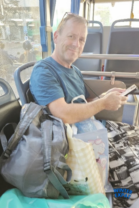 Z smiling sitting on a Bangkok public bus holding his phone, surrounded by shopping bags and a backpack after a day at the market. Original photo by Ariana of WinterExpat.