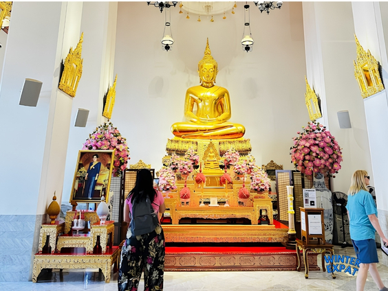 Golden seated Buddha inside one of Wat Pho's temple halls, decorated with floral arrangements and visited by tourists in Bangkok, Thailand. Original photo by Ariana of WinterExpat.