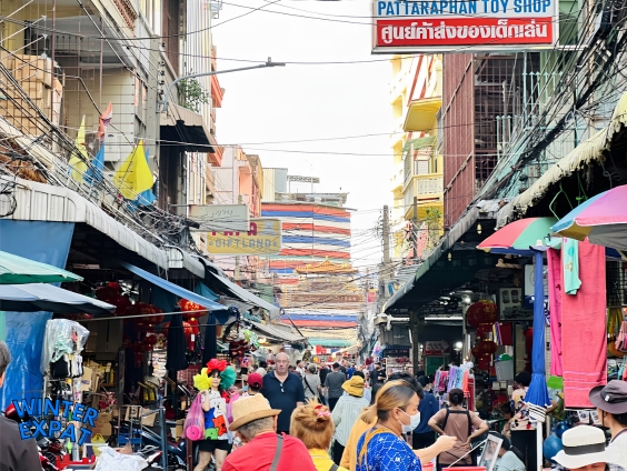 Crowded street view looking down a lane at Sampheng Market with awnings, hanging merchandise, and store signage overhead. Original photo by Ariana of WinterExpat.