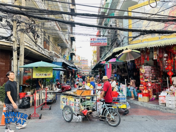 Busy outdoor entrance to Sampheng Market in Bangkok with street vendors, food carts, and shop stalls under overhead wires. Original photo by Ariana of WinterExpat.